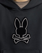 A close-up of the KIDS ALEXANDER HOODIE - B0H154E200 by Psycho Bunny reveals an embroidered rabbit's head with pointed ears above crossed bones, all beautifully stitched with a silver outline. This versatile black pullover hoodie offers a comfortable relaxed fit, perfect for any adventure.