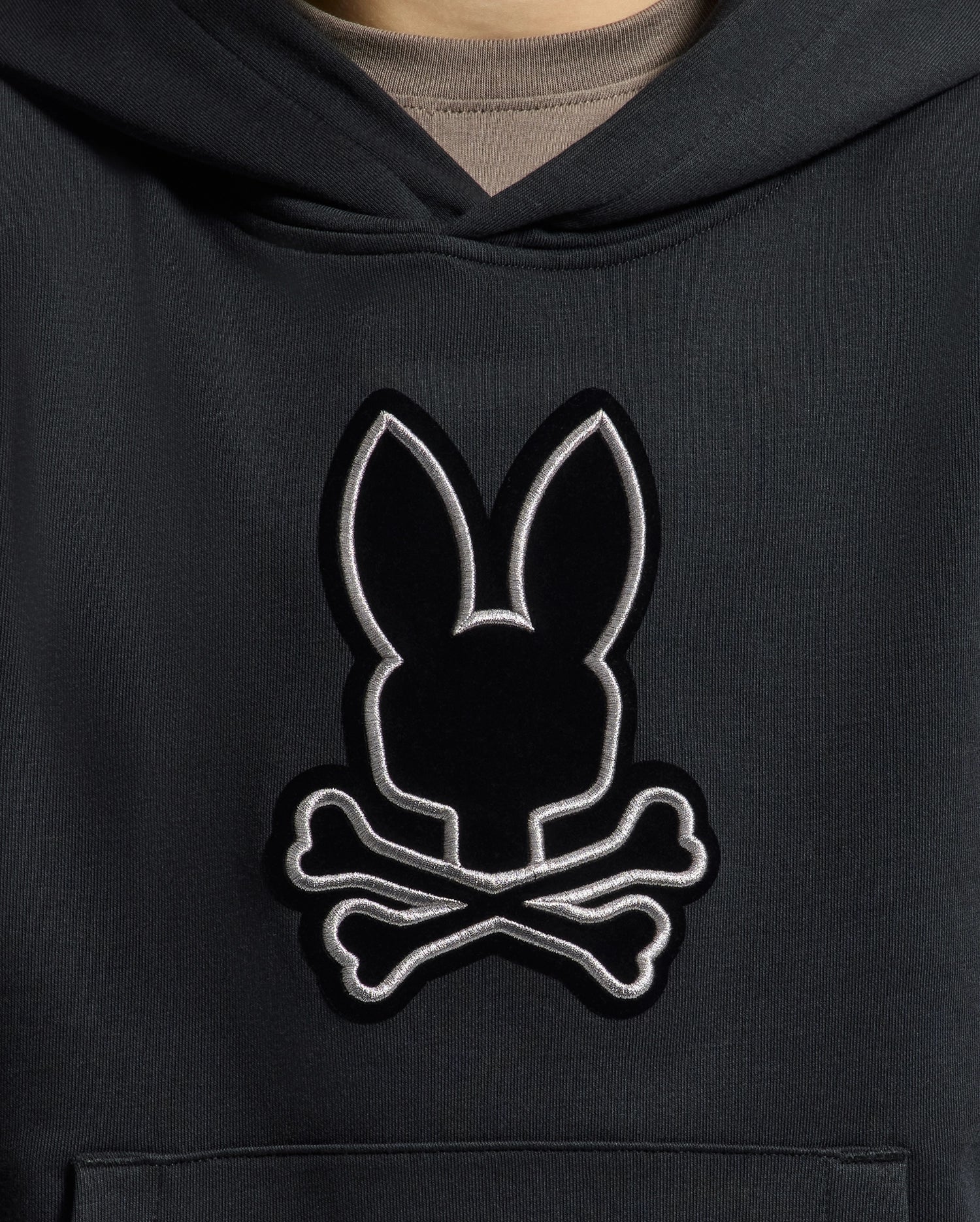 A close-up of the KIDS ALEXANDER HOODIE - B0H154E200 by Psycho Bunny reveals an embroidered rabbit's head with pointed ears above crossed bones, all beautifully stitched with a silver outline. This versatile black pullover hoodie offers a comfortable relaxed fit, perfect for any adventure.