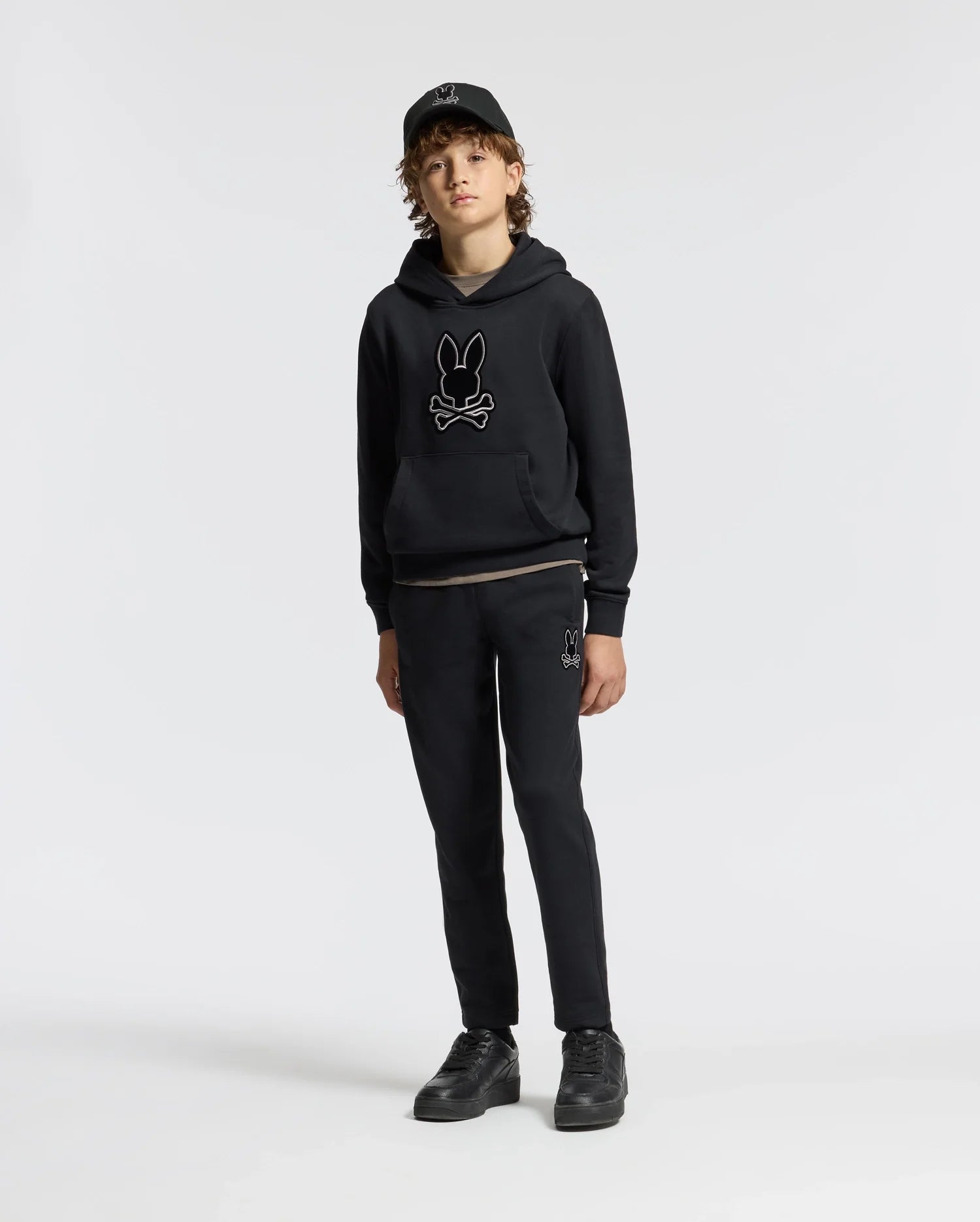 A person with curly hair stands facing forward, wearing the Psycho Bunny KIDS ALEXANDER HOODIE - B0H154E200 in black. This versatile pullover hoodie, featuring a bunny design and a comfortable relaxed fit, is paired with black pants and a black cap, standing out against the plain white background.