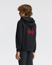 A person with curly hair is wearing the Psycho Bunny KIDS PIERCE ESSENTIAL FRENCH TERRY ZIP HOODIE (B0H269E200), which features an embroidered red skull and crossbones design on the back. The black hoodie combines coziness and style, crafted from soft French terry. Facing away from the camera, the individual stands against a plain, light-colored background.