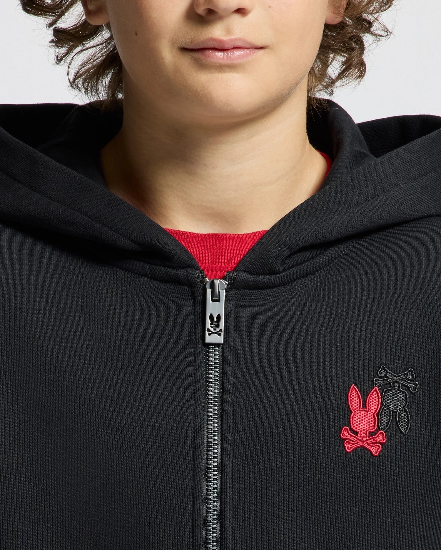 A person is wearing a black Kids Pierce Essential French Terry Zip Hoodie by Psycho Bunny, featuring a red and black bunny skull emblem on the chest. The zipper is partially closed, subtly revealing the comfort of the French Terry fabric. The focus remains on the hoodie design, as their face is partly obscured.