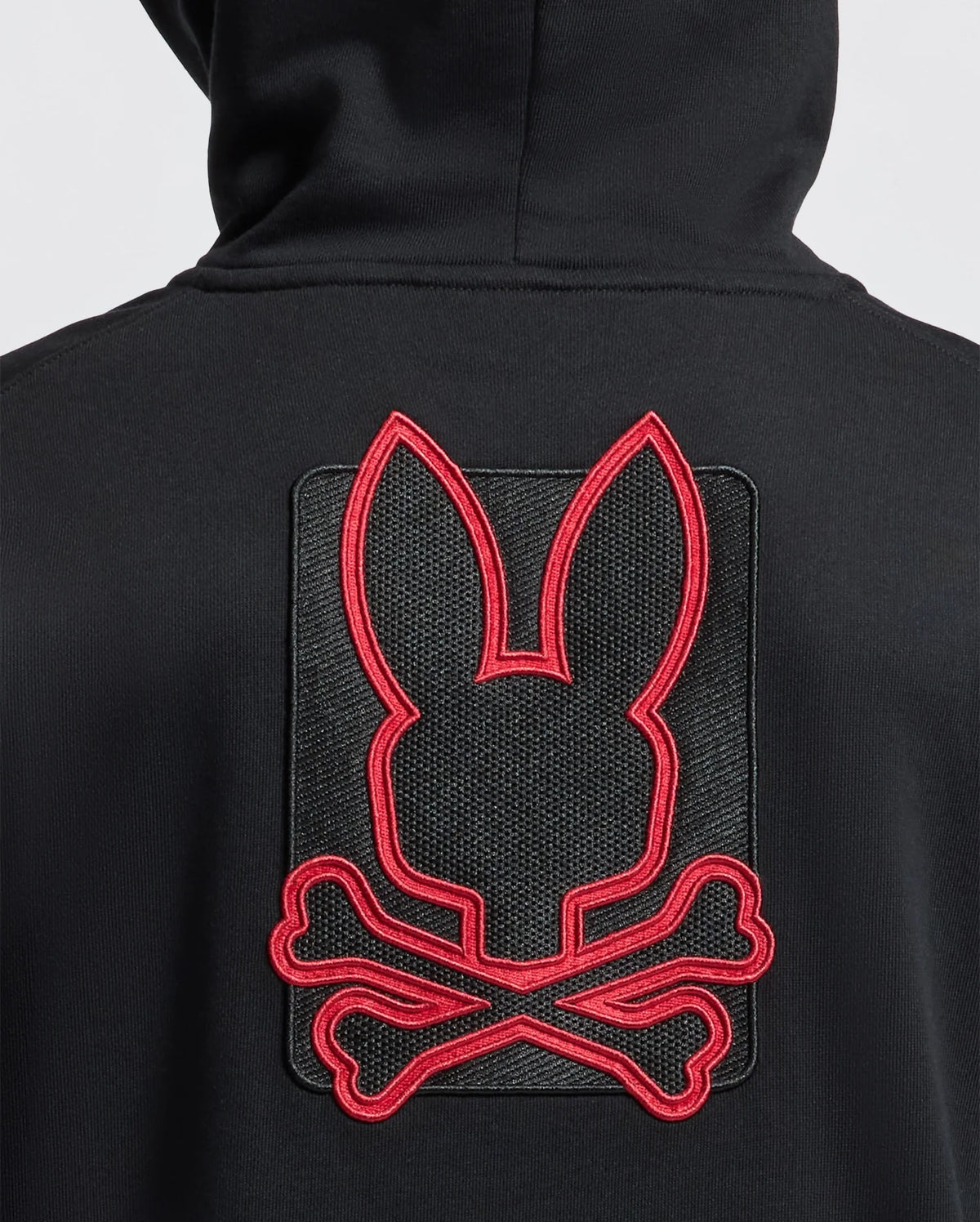 A person wearing the KIDS PIERCE ESSENTIAL FRENCH TERRY ZIP HOODIE by Psycho Bunny, which showcases a large red outline of a bunny head above crossed bones on the back. The unique emblem merges elements of a skull and crossbones with bunny ears, bringing an unexpected twist to this classic style.