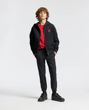 A person with curly hair stands confidently, wearing a black KIDS PIERCE ESSENTIAL FRENCH TERRY ZIP HOODIE by Psycho Bunny, featuring a red emblem. They pair it with a matching red shirt, black jogger pants, and black sneakers. The minimalist background enhances their bold style.