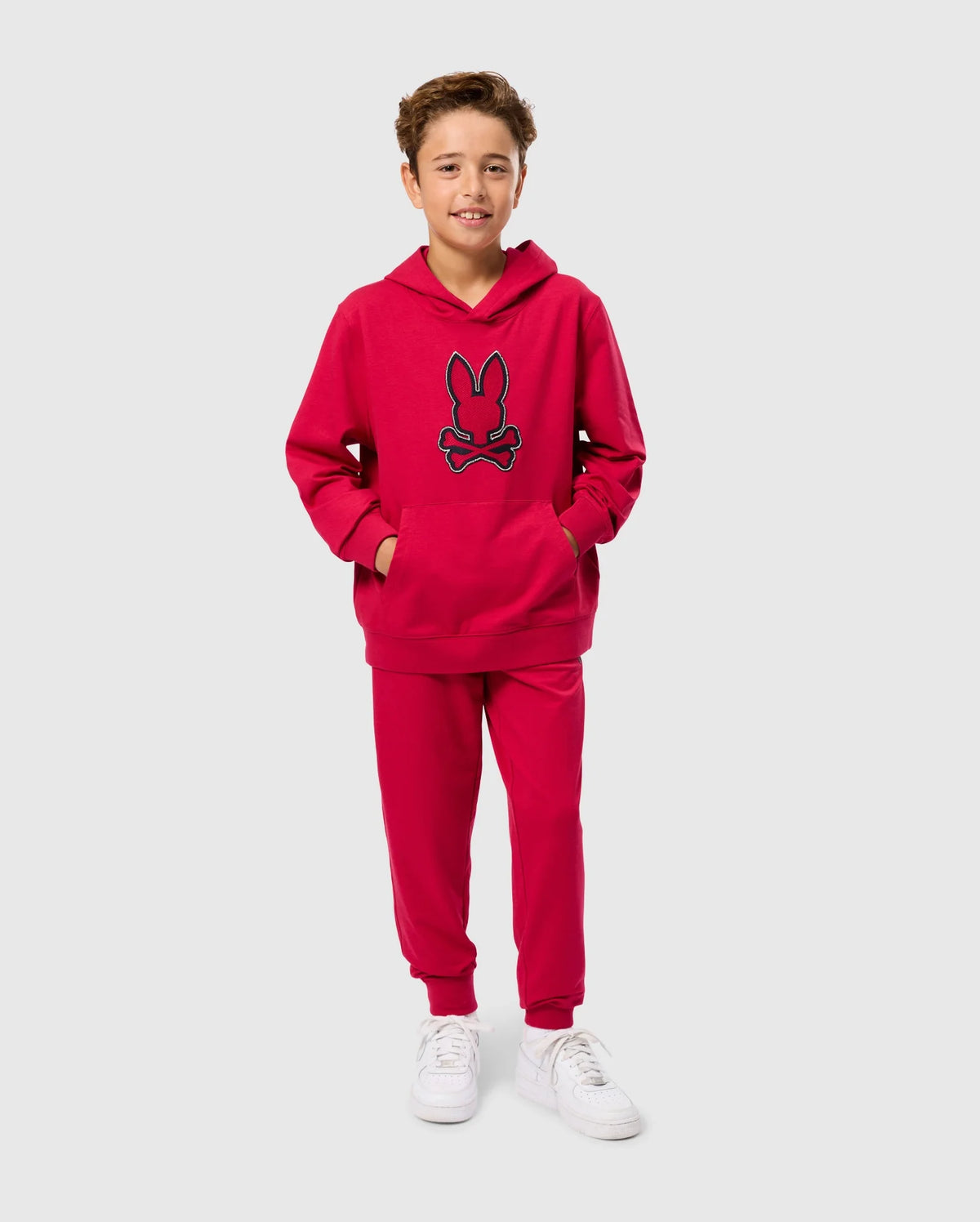 A young boy stands smiling, wearing the Psycho Bunny KIDS WALTER LIGHTWEIGHT HOODIE - B0H476D200 in bright red Micro French terry fabric that features a blue-outlined rabbit face design, along with matching red sweatpants. He has his hands in the sweatshirt pockets and is also wearing white sneakers. The background is plain light gray.