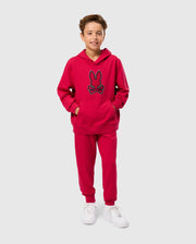 A young boy stands smiling, wearing the Psycho Bunny KIDS WALTER LIGHTWEIGHT HOODIE - B0H476D200 in bright red Micro French terry fabric that features a blue-outlined rabbit face design, along with matching red sweatpants. He has his hands in the sweatshirt pockets and is also wearing white sneakers. The background is plain light gray.