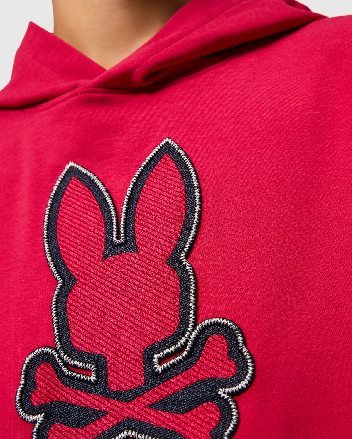 Close-up image of a person wearing a red KIDS WALTER LIGHTWEIGHT HOODIE - B0H476D200 by Psycho Bunny, featuring a stitched design of a bunny's head with crossed bones underneath. The design is predominantly black with red accents, and the surrounding stitching is white. This piece adds a modern twist to casual wear. The person's face is not visible.