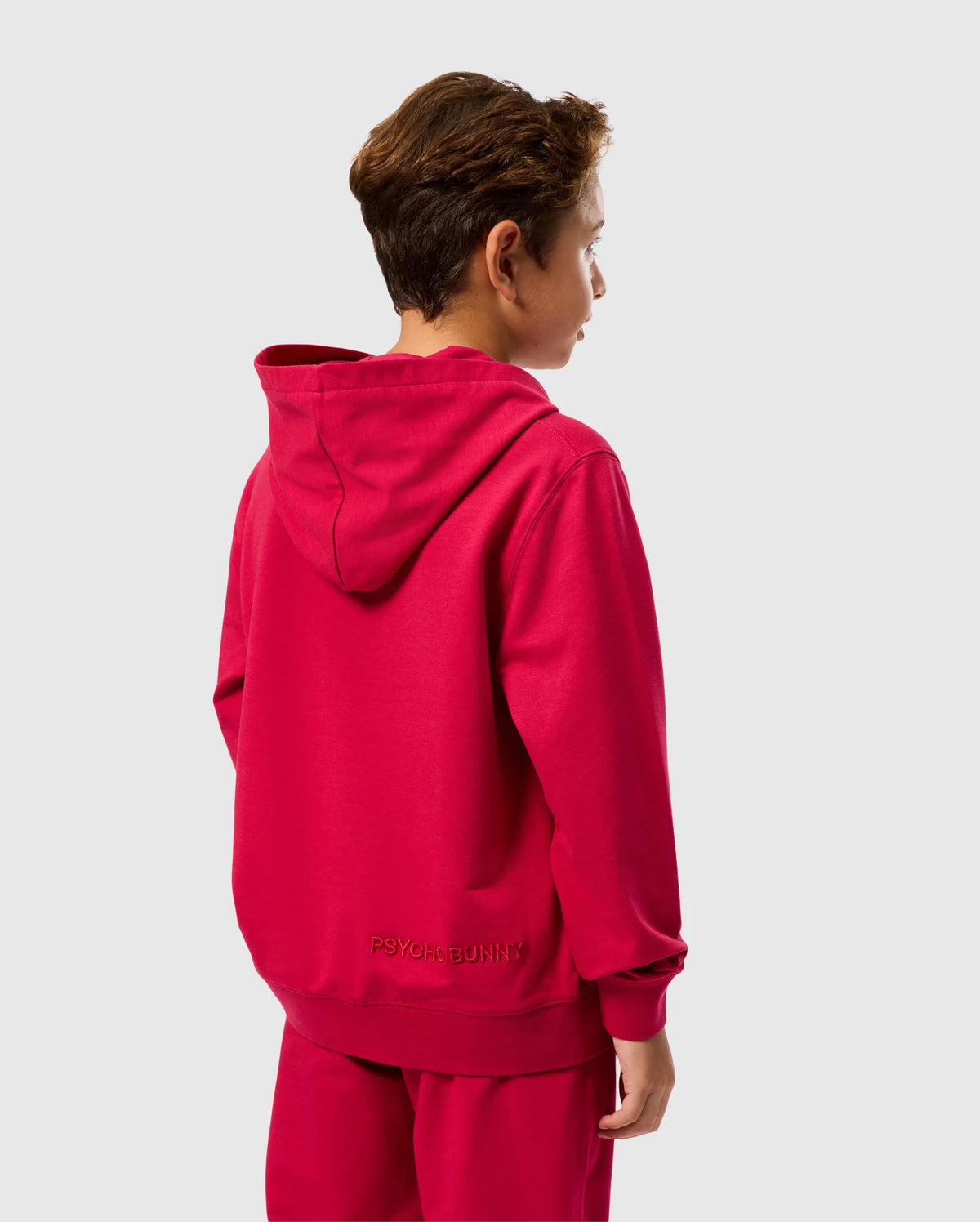 A person with short brown hair is seen from behind, wearing the KIDS WALTER LIGHTWEIGHT HOODIE in bright red made of Micro French terry, along with matching pants. The back of the hoodie features the text "PSYCHO BUNNY" near the bottom, giving it a modern twist. The background is plain white.