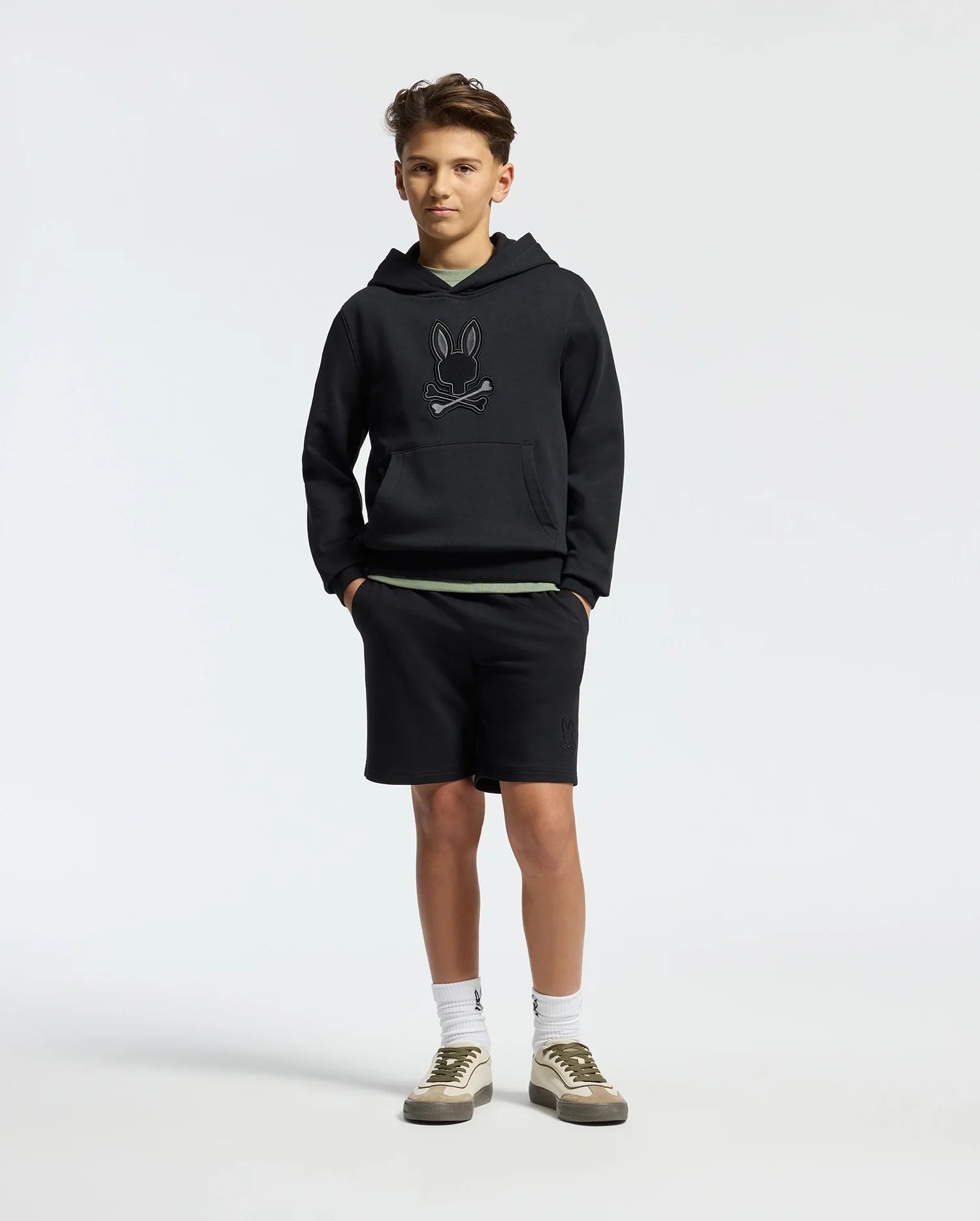A teenage boy wears a Psycho Bunny KIDS NEW CASTLE FRENCH TERRY HOODIE, showcasing its embroidered Bunny logo. He pairs it with black shorts, white socks, and white sneakers featuring olive accents while standing casually with hands in pockets against a plain white background.