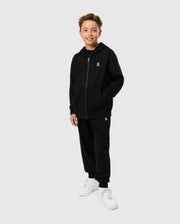 A smiling boy stands against a plain light gray background, wearing a black Psycho Bunny KIDS LEVY ZIP FLEECE HOODIE - B0H720D200 and matching black sweatpants from the kids' essential collection. He has one hand in his pocket and is wearing white sneakers. The hoodie and sweatpants each feature a small embroidered logo on them.