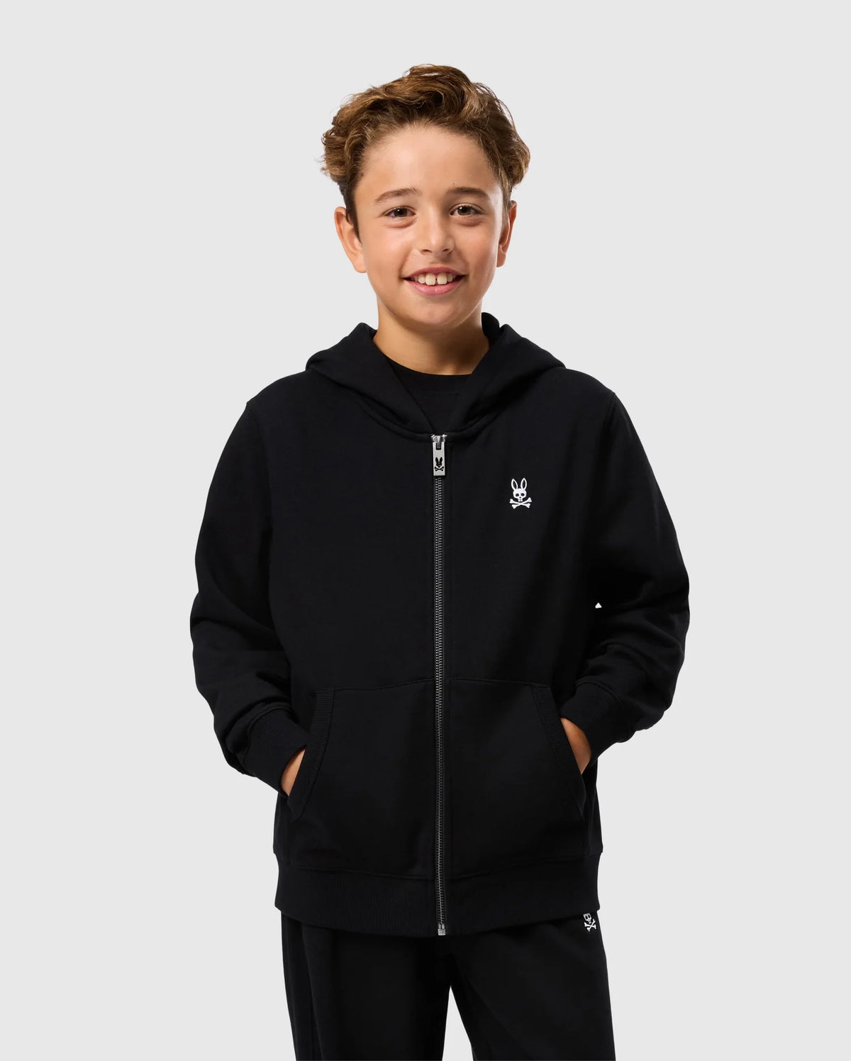A smiling young boy stands against a plain background, wearing a Psycho Bunny KIDS LEVY ZIP FLEECE HOODIE - B0H720D200 in black with his hands in the pockets and dark pants. This essential hoodie for kids, made from 100% cotton, features a small white skull and crossbones logo on the left chest.