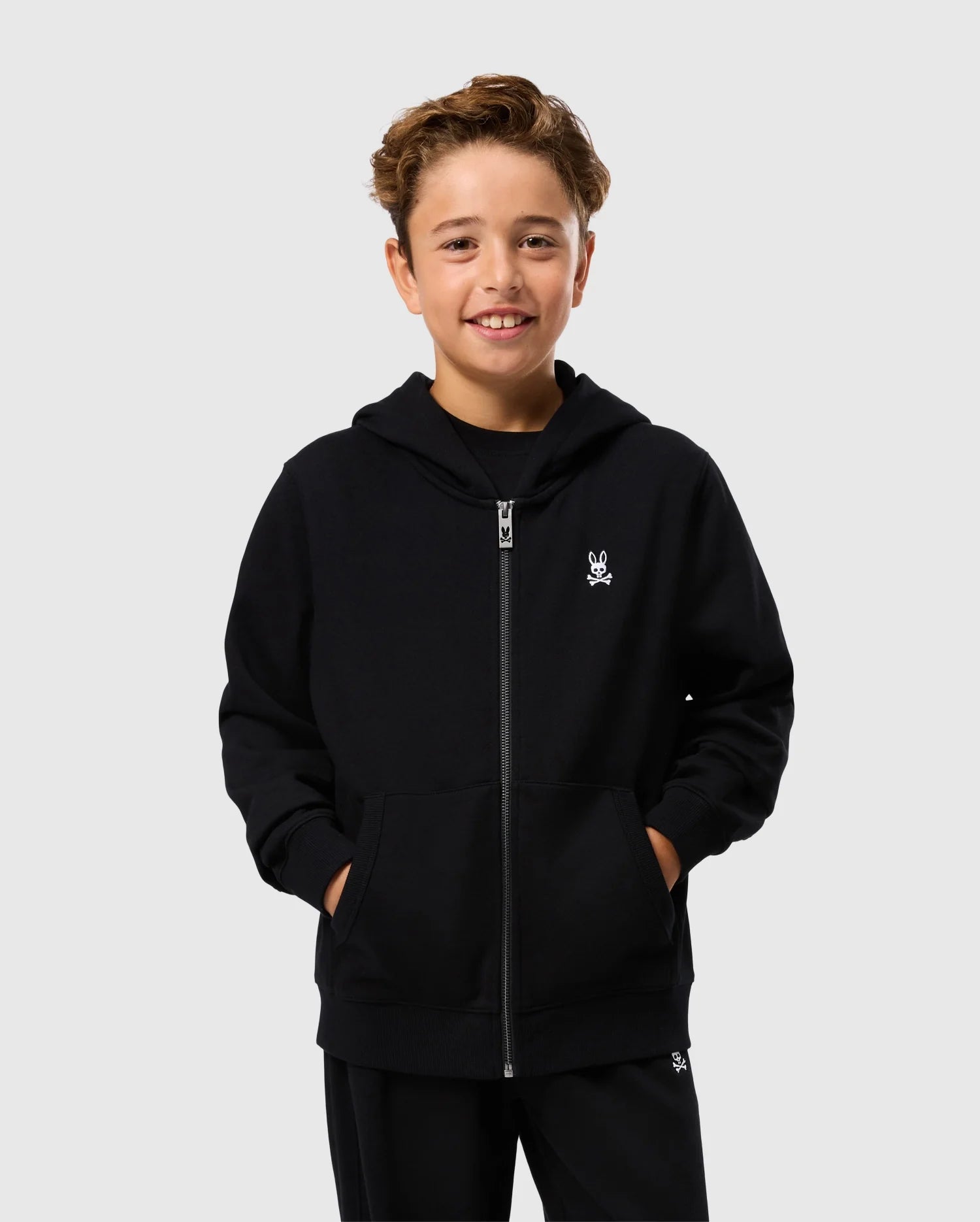 A smiling young boy stands against a plain background, wearing a Psycho Bunny KIDS LEVY ZIP FLEECE HOODIE - B0H720D200 in black with his hands in the pockets and dark pants. This essential hoodie for kids, made from 100% cotton, features a small white skull and crossbones logo on the left chest.