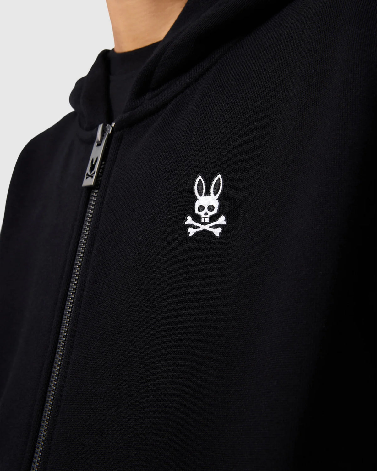 Close-up of the Psycho Bunny KIDS LEVY ZIP FLEECE HOODIE - B0H720D200, a black essential French terry hoodie with a zipper and a white embroidered logo on the chest. The logo features a bunny head with long ears above a pair of crossed bones. The image focuses on the upper half of the 100% cotton hoodie, including part of the zipper and hood.