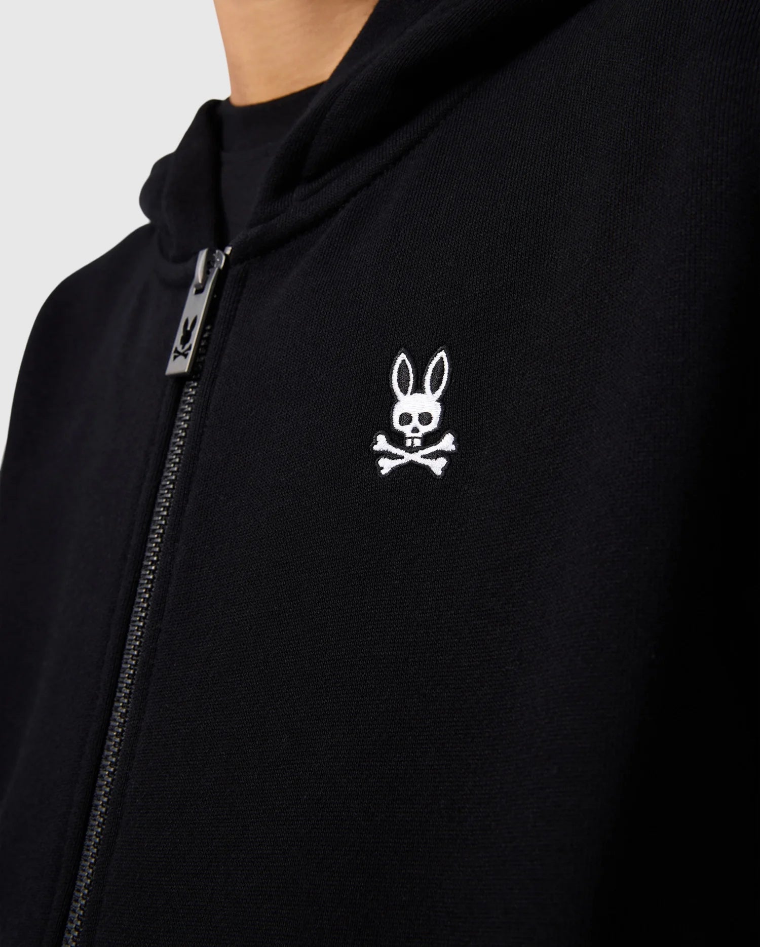 Close-up of the Psycho Bunny KIDS LEVY ZIP FLEECE HOODIE - B0H720D200, a black essential French terry hoodie with a zipper and a white embroidered logo on the chest. The logo features a bunny head with long ears above a pair of crossed bones. The image focuses on the upper half of the 100% cotton hoodie, including part of the zipper and hood.