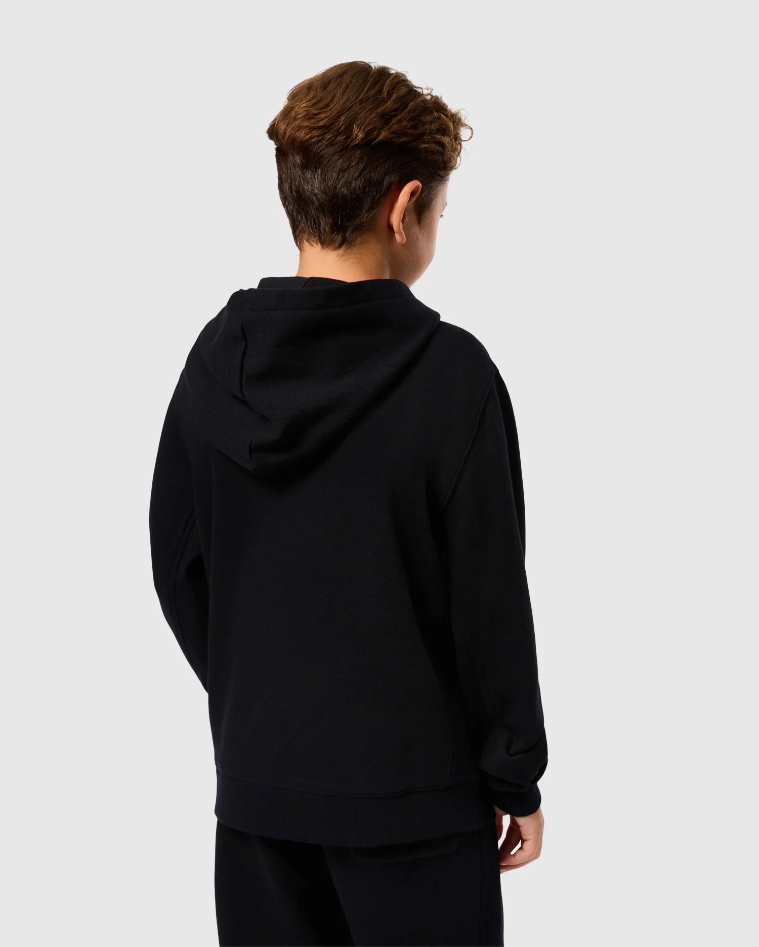 A person with short, wavy hair is shown from behind, wearing the Psycho Bunny KIDS LEVY ZIP FLEECE HOODIE - B0H720D200 and black pants. They are standing against a plain, light-colored background.