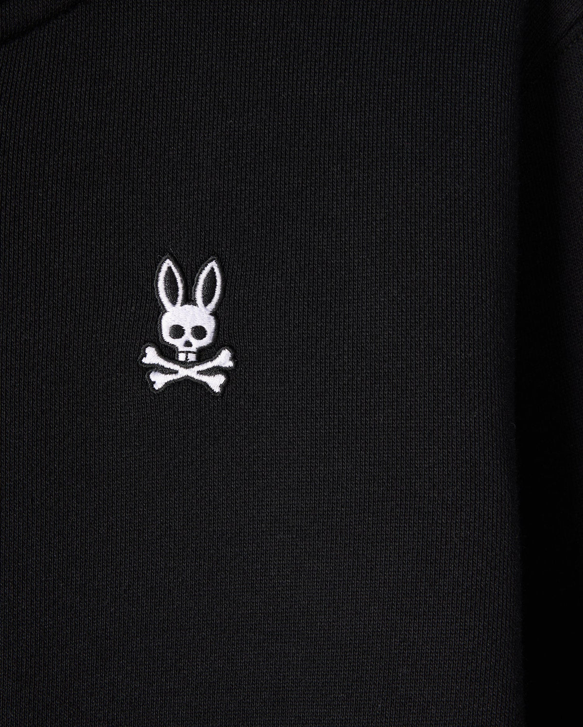 A close-up of the Psycho Bunny KIDS LEVY ZIP FLEECE HOODIE - B0H720D200, featuring a black color with a white embroidered bunny logo. The logo presents a long-eared bunny head with a skull face, above crossed bones, offering a playful and edgy twist on the traditional skull and crossbones symbol.