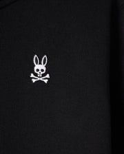 A close-up of the Psycho Bunny KIDS LEVY ZIP FLEECE HOODIE - B0H720D200, featuring a black color with a white embroidered bunny logo. The logo presents a long-eared bunny head with a skull face, above crossed bones, offering a playful and edgy twist on the traditional skull and crossbones symbol.