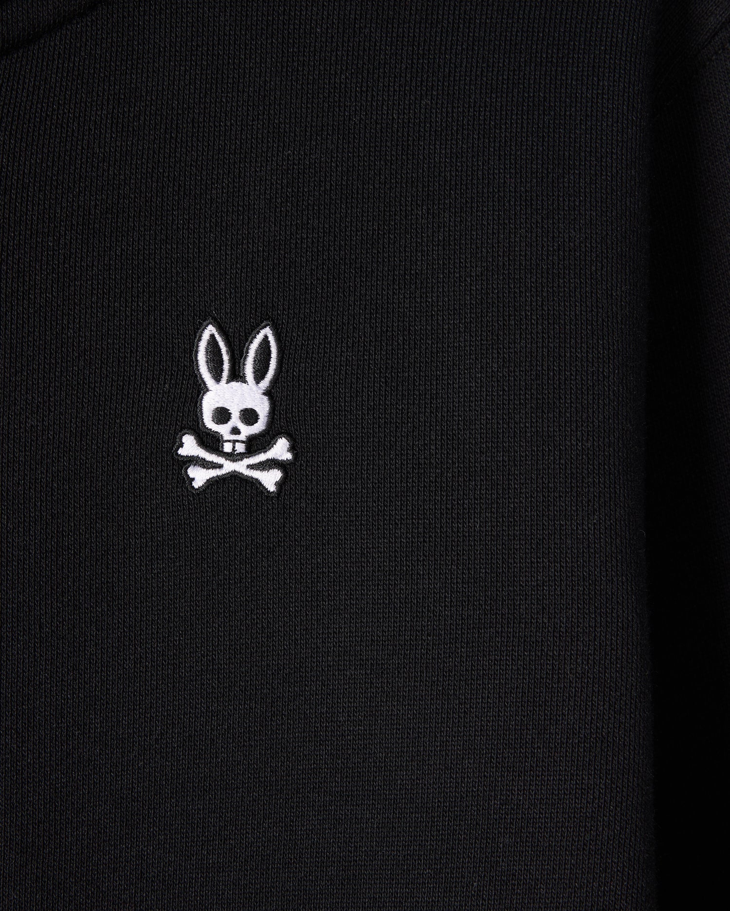 A close-up of the Psycho Bunny KIDS LEVY ZIP FLEECE HOODIE - B0H720D200, featuring a black color with a white embroidered bunny logo. The logo presents a long-eared bunny head with a skull face, above crossed bones, offering a playful and edgy twist on the traditional skull and crossbones symbol.