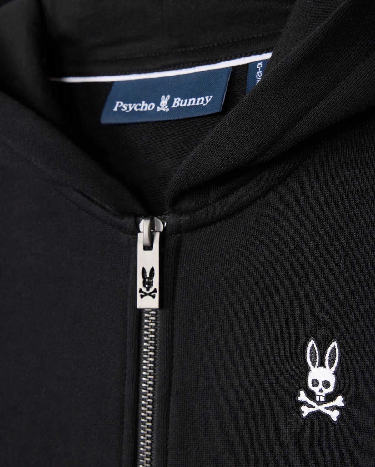 Close-up of the KIDS LEVY ZIP FLEECE HOODIE - B0H720D200 in black, highlighting a silver zipper and the distinctive bunny skull and crossbones logo on the chest. The label inside this 100% cotton hoodie features the brand name "Psycho Bunny." Perfectly blending comfort with edgy style, this piece is an essential addition to any kid's wardrobe.