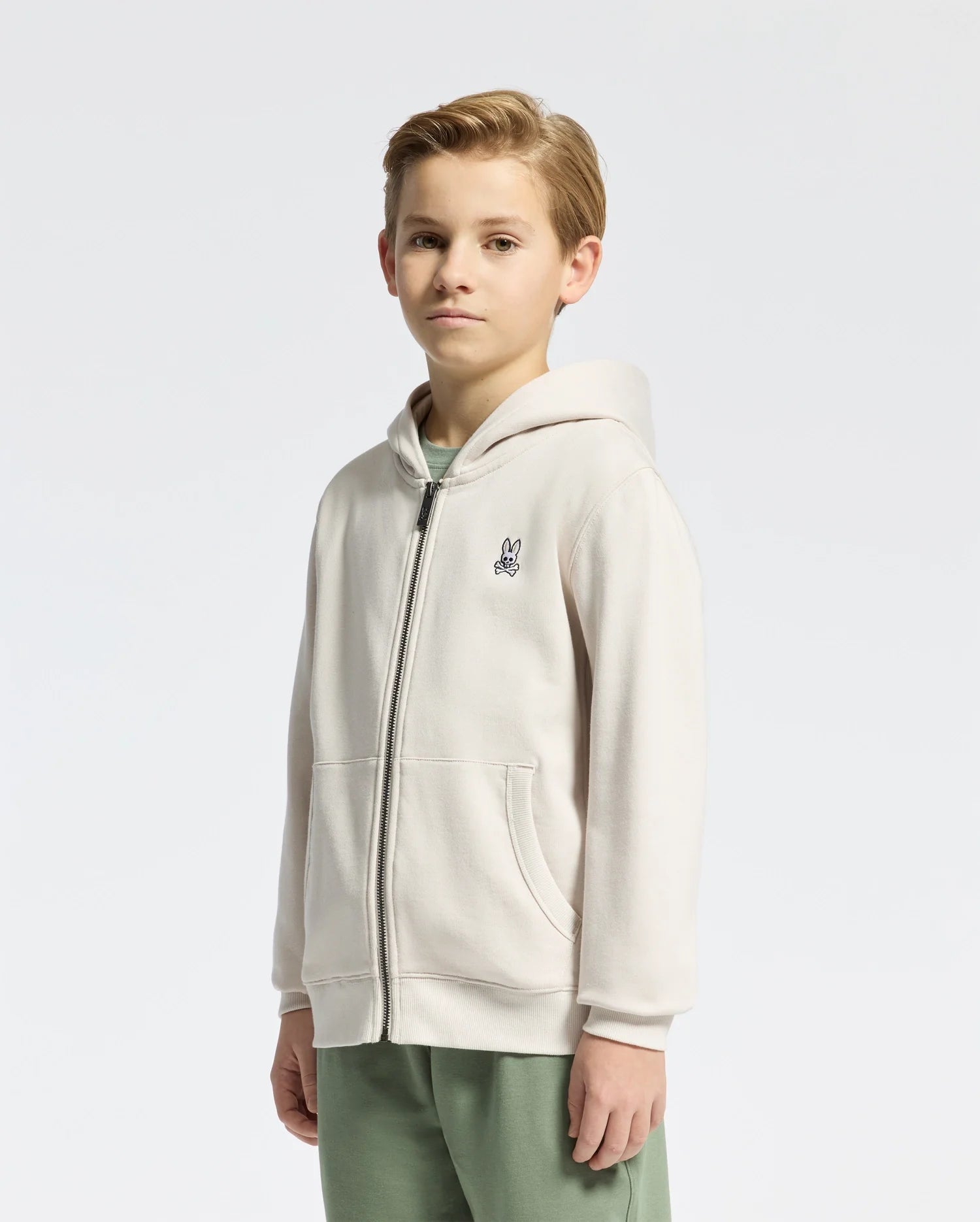 A young boy with short blond hair smiles slightly, wearing a light beige KIDS LEVY ESSENTIAL ZIP FLEECE HOODIE from Psycho Bunny, featuring a small embroidered logo on the chest, paired with green pants. Crafted from soft cotton for kids' comfort against a simple background.