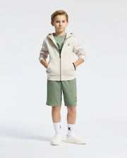 A boy stands confidently in a Psycho Bunny KIDS LEVY ESSENTIAL ZIP FLEECE HOODIE - B0H720D200, crafted from soft French terry. The light zip-up hoodie complements his green shirt and matching shorts. With hands in pockets, he sports white sneakers and socks featuring a graphic, set against a plain white background.
