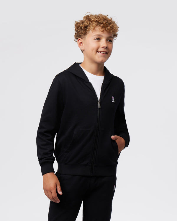 A smiling young boy with curly hair wearing a black Psycho Bunny KIDS CLASSIC FRENCH TERRY ZIP HOODIE - B0H826ARFT and matching pants, standing against a plain white background. He has his hand in a kangaroo pocket.