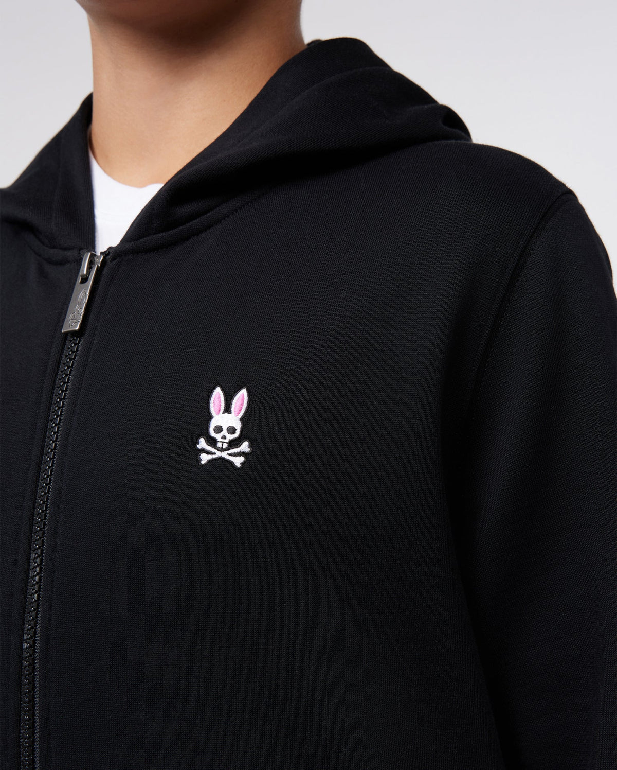 Close-up of a person wearing a black Psycho Bunny KIDS CLASSIC FRENCH TERRY ZIP HOODIE - B0H826ARFT with a small white and pink embroidered Bunny logo on the left chest area.