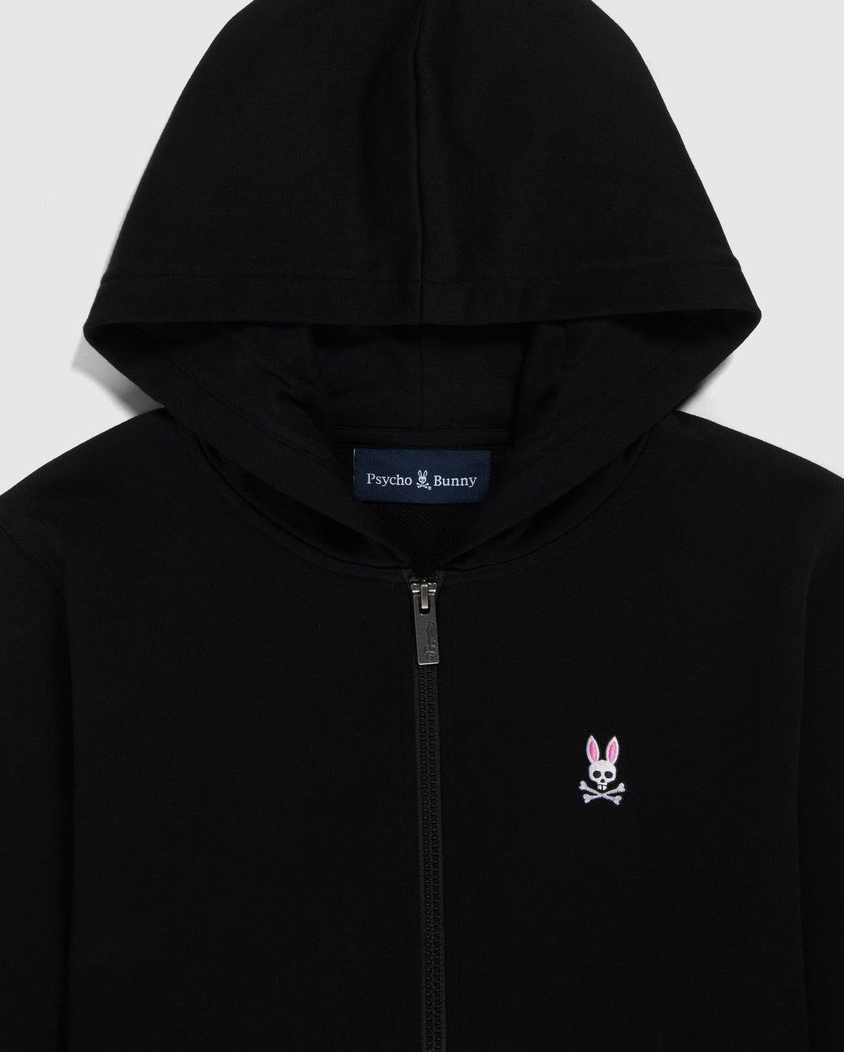 Close-up of a Psycho Bunny French terry zip hoodie in black with a silver zipper, featuring an embroidered Bunny logo on the left chest and the brand name 'Psycho Bunny' printed inside the collar.