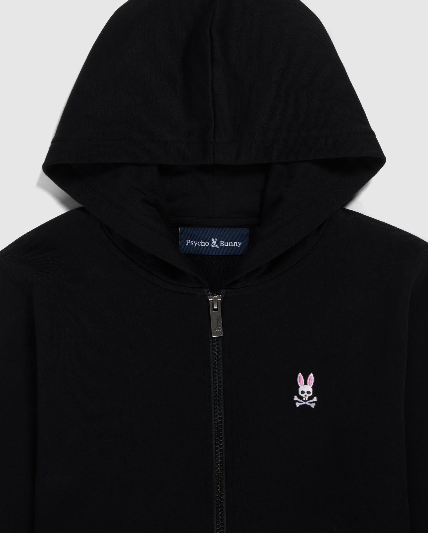 Close-up of a Psycho Bunny French terry zip hoodie in black with a silver zipper, featuring an embroidered Bunny logo on the left chest and the brand name 'Psycho Bunny' printed inside the collar.