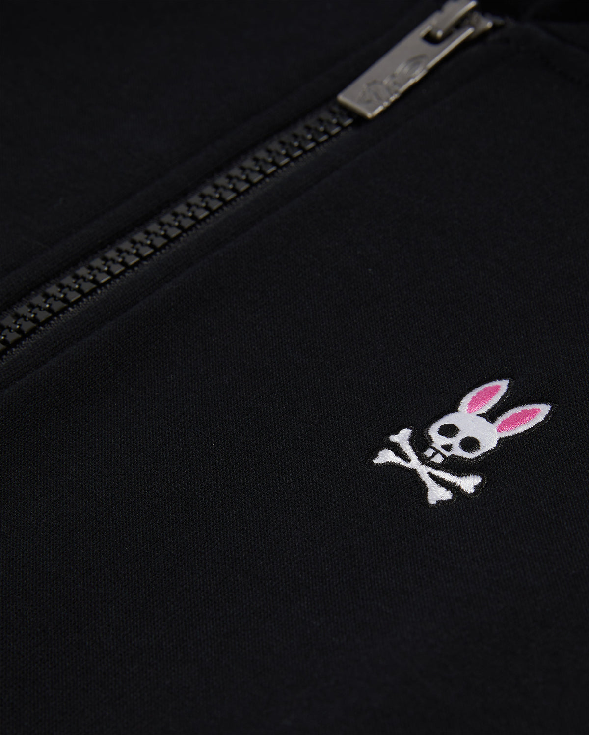 Close-up of a black Psycho Bunny KIDS CLASSIC FRENCH TERRY ZIP HOODIE with an embroidered bunny logo featuring a skull with crossed bones and pink ears, next to a partially zipped silver zipper.