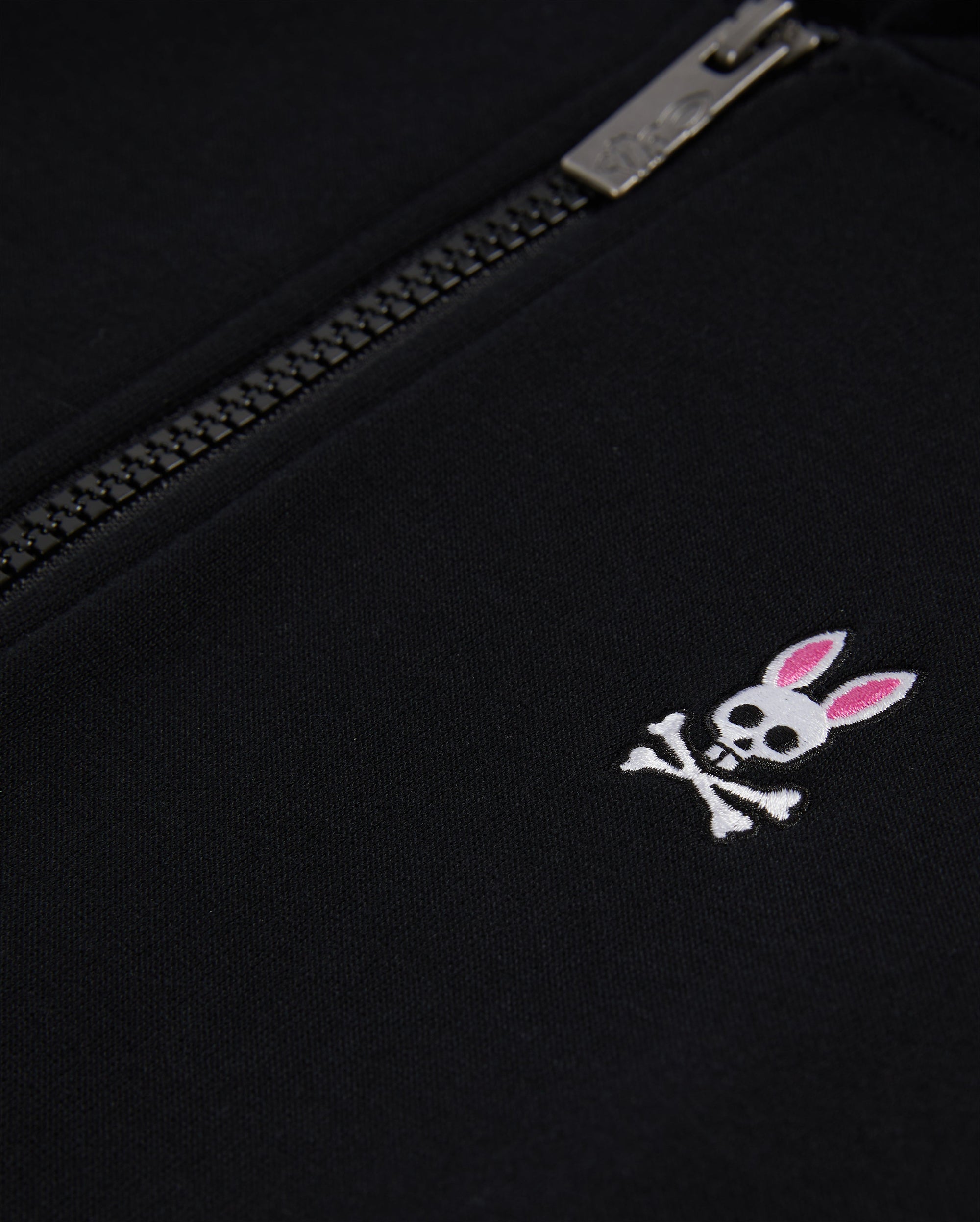 Close-up of a black Psycho Bunny KIDS CLASSIC FRENCH TERRY ZIP HOODIE with an embroidered bunny logo featuring a skull with crossed bones and pink ears, next to a partially zipped silver zipper.