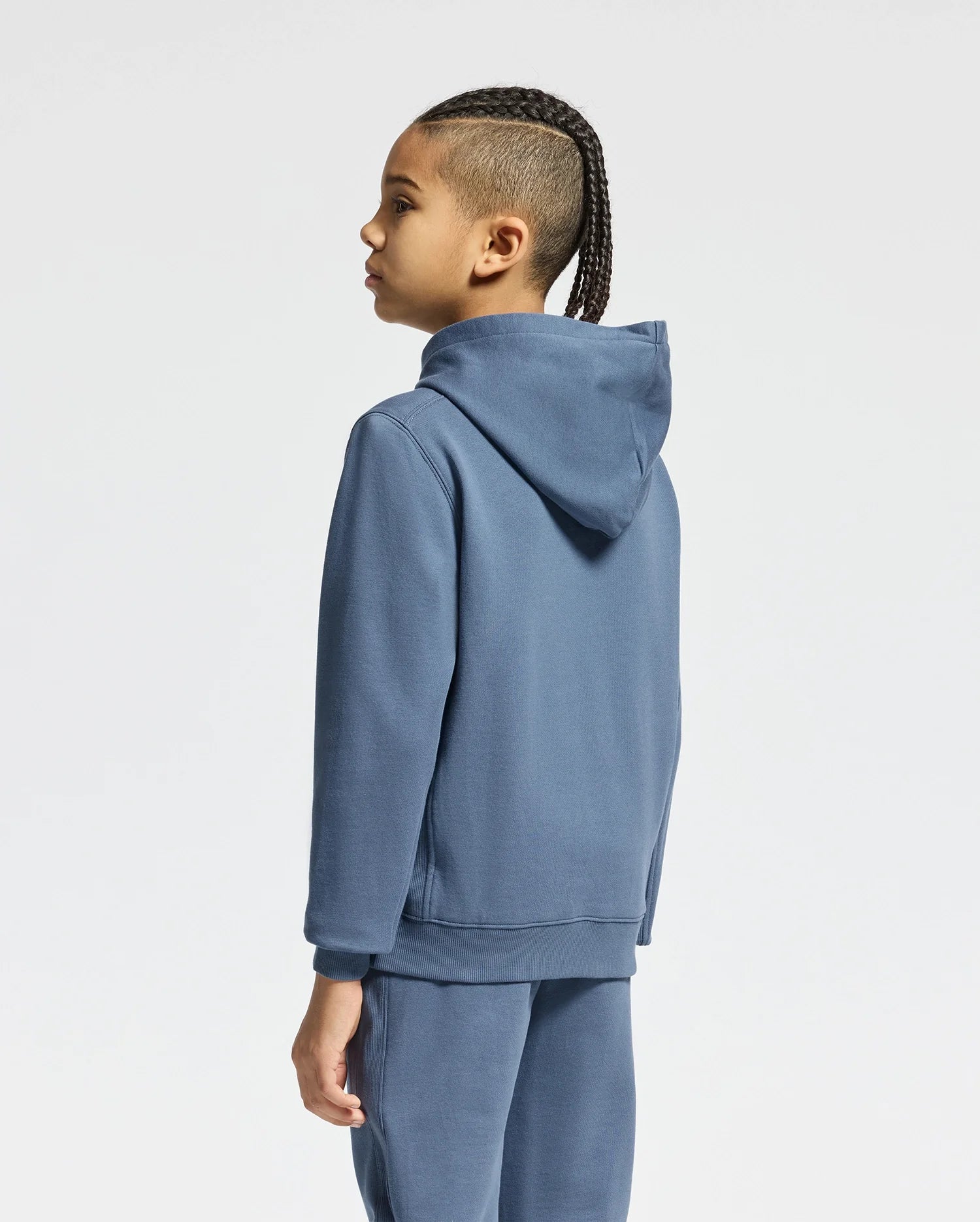 A person with braided hair stands facing away, wearing a Psycho Bunny KIDS Levy Essential Fleece Hoodie and matching fleece sweatpants in blue against a plain white background.