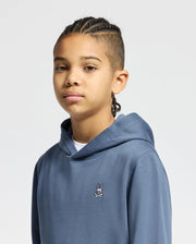 A young person with braided hair is wearing a Psycho Bunny KIDS LEVY ESSENTIAL FLEECE HOODIE - B0H859D200. This hoodie is crafted from 100% cotton and features a small cartoon logo on the chest, complementing the minimalist aesthetic of the plain light gray background.