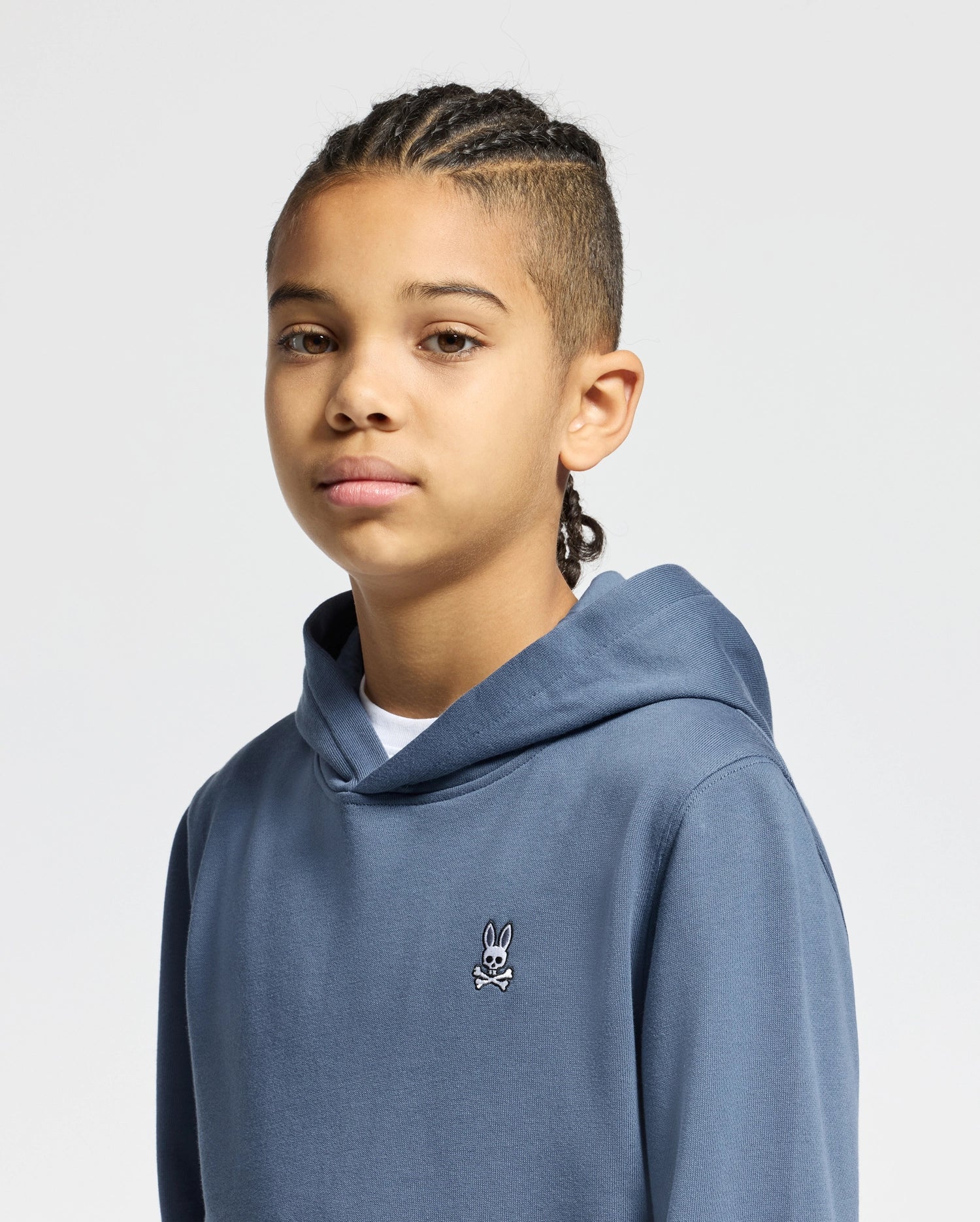 A young person with braided hair is wearing a Psycho Bunny KIDS LEVY ESSENTIAL FLEECE HOODIE - B0H859D200. This hoodie is crafted from 100% cotton and features a small cartoon logo on the chest, complementing the minimalist aesthetic of the plain light gray background.