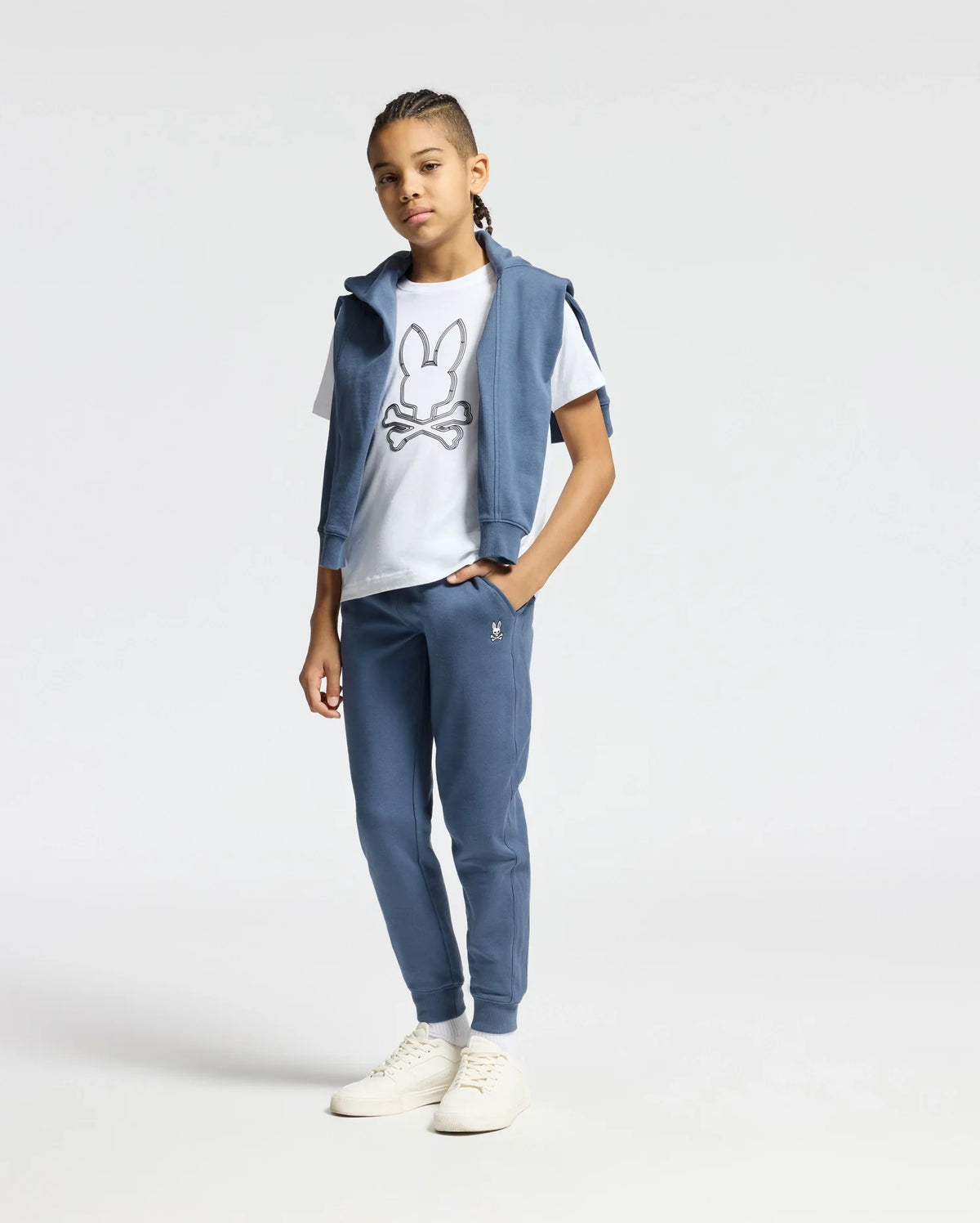 A young person with braided hair stands wearing a blue tracksuit and a white T-shirt featuring a cartoon design. Enhancing their outfit are the Kids Levy Essential Fleece Hoodie by Psycho Bunny, fleece sweatpants, and white sneakers. The plain, light-colored background allows the ensemble to stand out effortlessly.