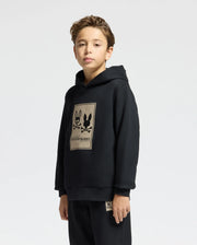 A young person in a black Psycho Bunny KIDS MONROE TWILL PATCH FLEECE HOODIE, featuring a graphic with two bunnies and crossbones on the front, stands against a plain white cotton backdrop. The individual has short brown hair.
