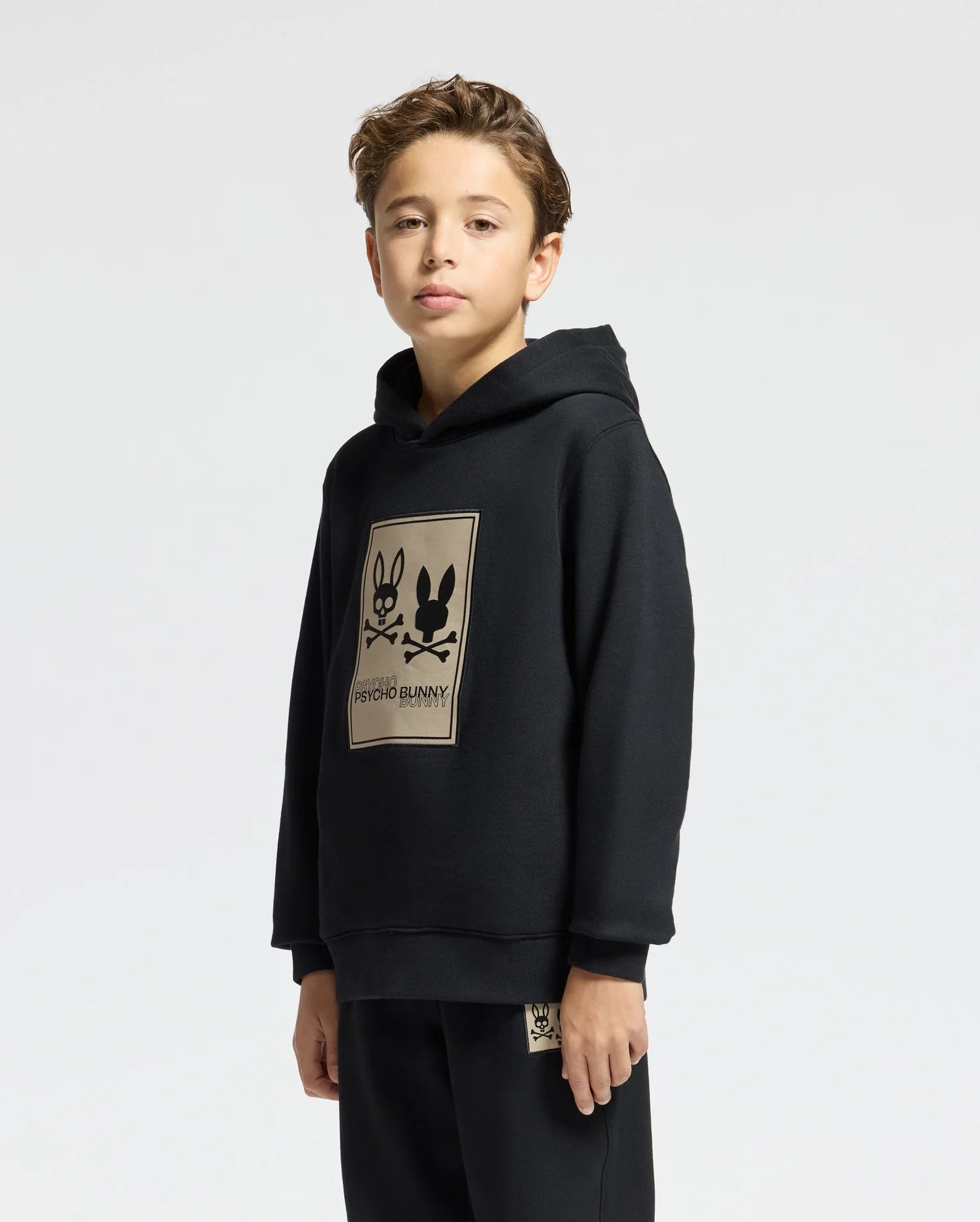 A young person in a black Psycho Bunny KIDS MONROE TWILL PATCH FLEECE HOODIE, featuring a graphic with two bunnies and crossbones on the front, stands against a plain white cotton backdrop. The individual has short brown hair.