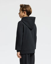 A young person with light brown hair stands sideways against a plain white background, wearing Psycho Bunny's black KIDS MONROE TWILL PATCH FLEECE HOODIE - B0H926D200 and matching pants. The face is not visible, showing only the side and back.