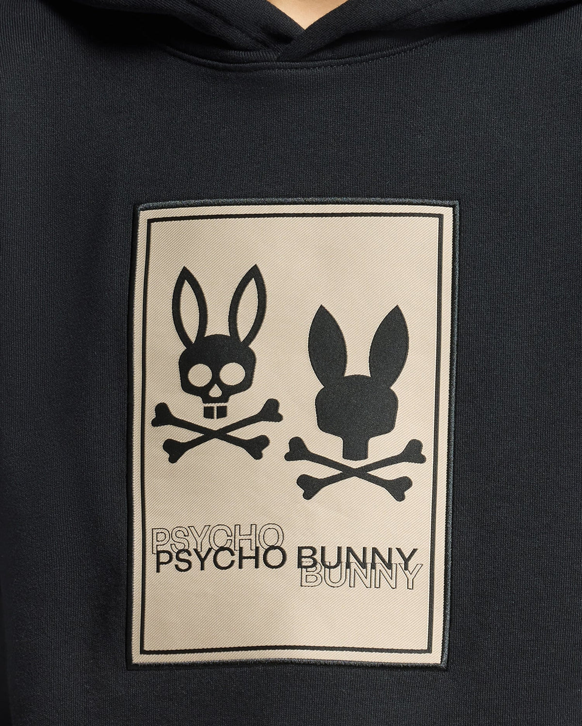 The KIDS MONROE TWILL PATCH FLEECE HOODIE by Psycho Bunny features a cozy French terry design with two black bunny heads—one with crossbones and a skull nose, the other simpler—above "Psycho Bunny" on a patch. Ideal for blending comfort and edgy style.
