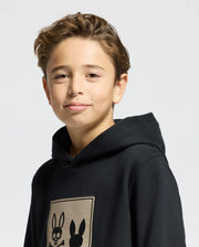 A young boy with short brown hair is wearing the Psycho Bunny KIDS MONROE TWILL PATCH FLEECE HOODIE - B0H926D200, featuring two bunny silhouettes with a skull design. He stands against a white background and smiles slightly at the camera.