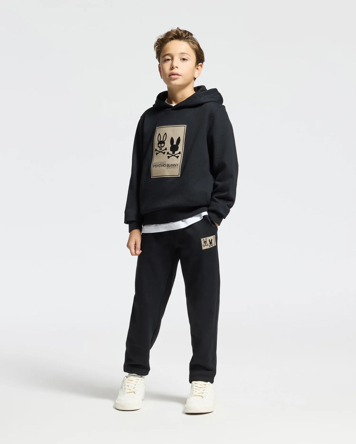 A young boy confidently wears the KIDS MONROE TWILL PATCH FLEECE HOODIE by Psycho Bunny, a black tracksuit with a cartoon design on the French terry hoodie and pants. He pairs it with white sneakers against a plain white background.