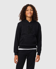 A young person with curly hair smiles while wearing a black Psycho Bunny KIDS PACIFIC CHENILLE HOODIE (B0H930D200) featuring a rabbit design in chenille embroidery. The background is plain light gray.