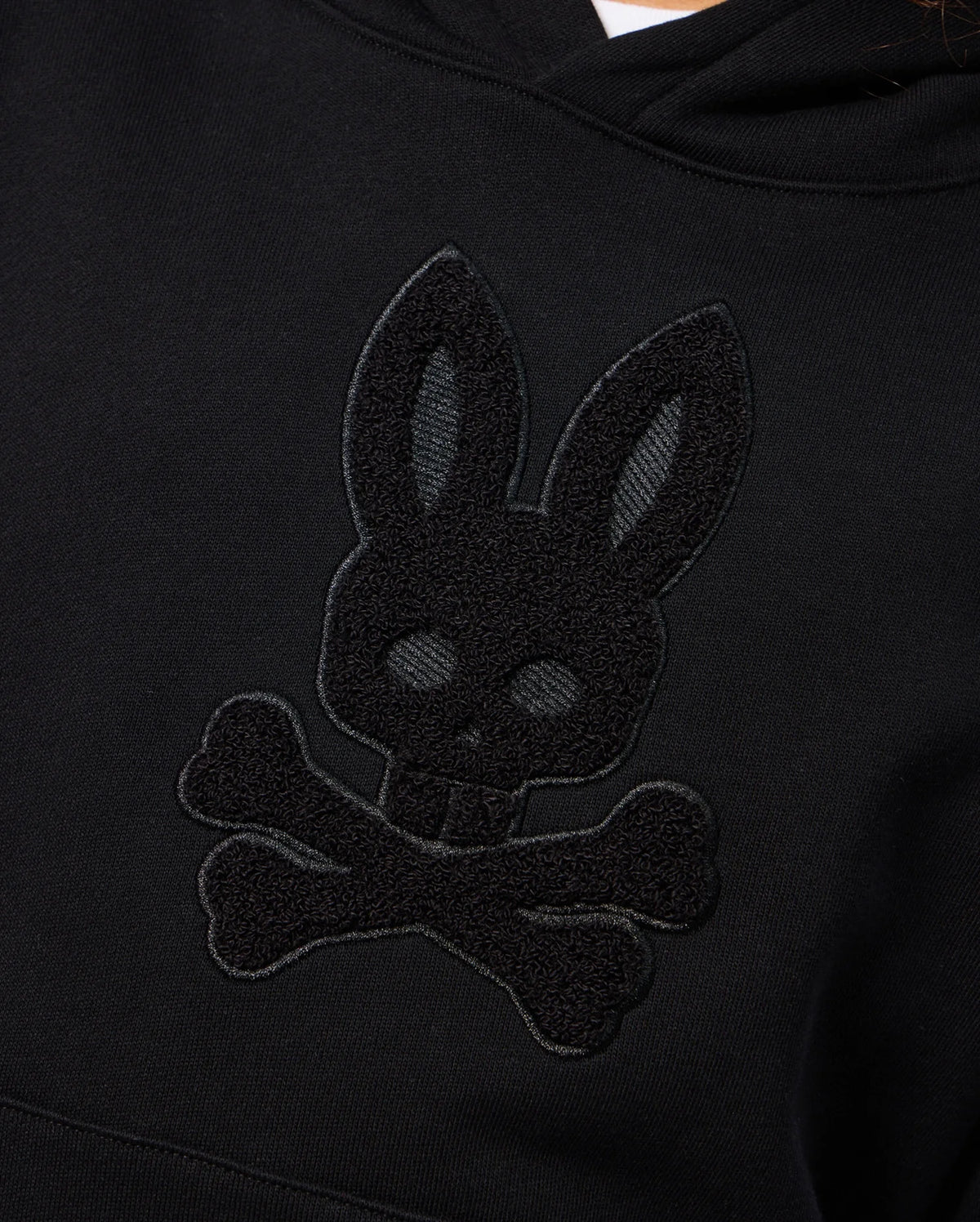 A detailed look at the KIDS PACIFIC CHENILLE HOODIE - B0H930D200 by Psycho Bunny, featuring a chenille embroidery of a bunny's head with skull-like features and crossed bones beneath it, resembling a stylized skull and crossbones symbol on a black French terry pullover hoodie.