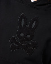 A detailed look at the KIDS PACIFIC CHENILLE HOODIE - B0H930D200 by Psycho Bunny, featuring a chenille embroidery of a bunny's head with skull-like features and crossed bones beneath it, resembling a stylized skull and crossbones symbol on a black French terry pullover hoodie.