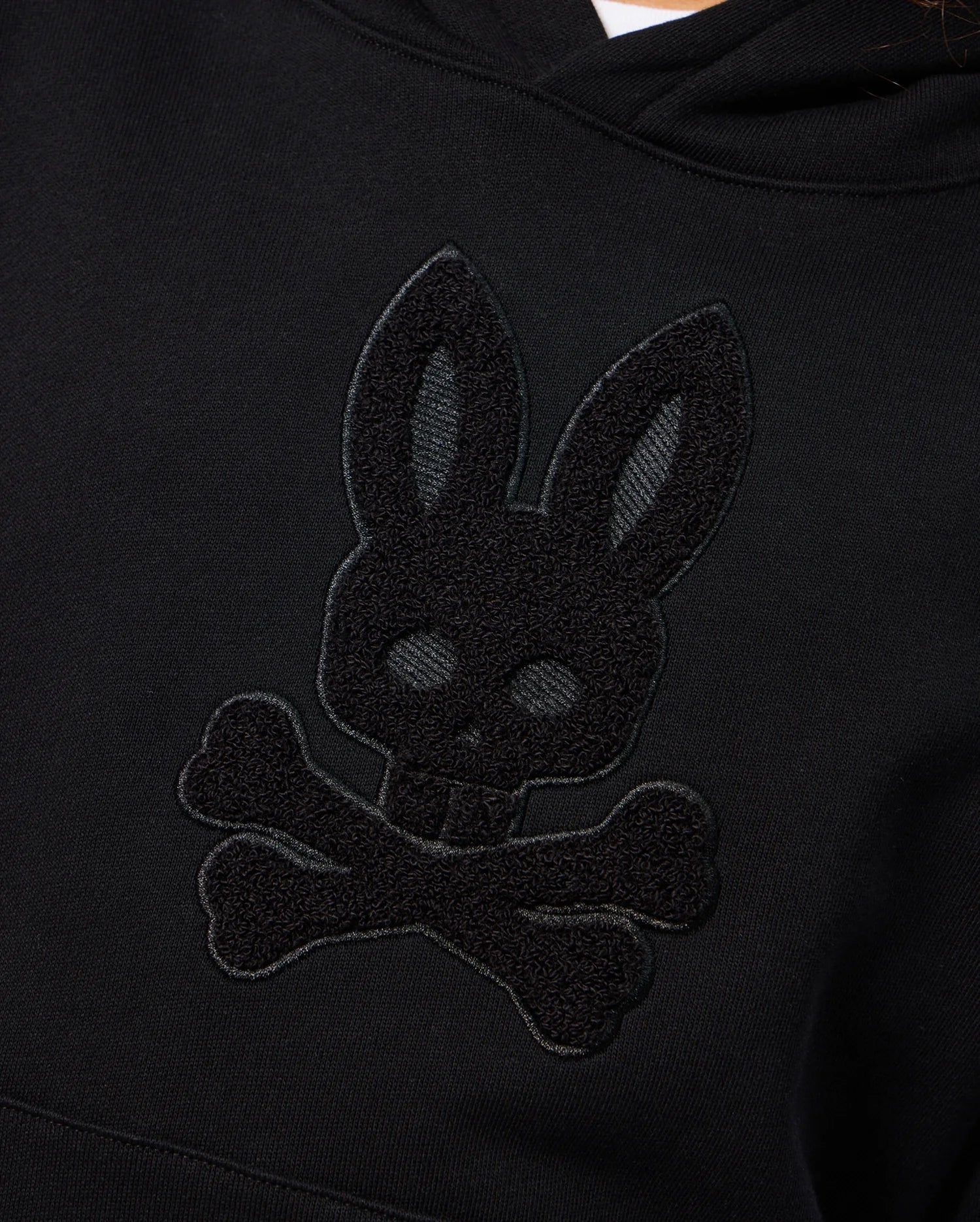 A detailed look at the KIDS PACIFIC CHENILLE HOODIE - B0H930D200 by Psycho Bunny, featuring a chenille embroidery of a bunny's head with skull-like features and crossed bones beneath it, resembling a stylized skull and crossbones symbol on a black French terry pullover hoodie.