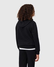 A person with curly hair, wearing a black KIDS PACIFIC CHENILLE HOODIE - B0H930D200 by Psycho Bunny and black pants, stands facing away from the camera against a gray background.