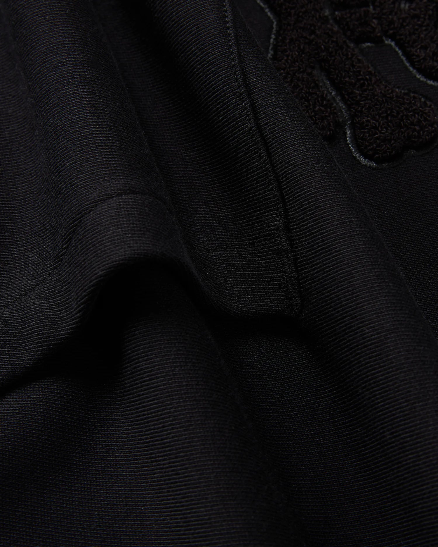 Here's a close-up of the black textured fabric from the KIDS PACIFIC CHENILLE HOODIE by Psycho Bunny (B0H930D200). The visible seam and part of the raised chenille embroidery design stand out. The soft fabric is slightly folded, emphasizing stitching details that evoke the cozy feel of a French terry hoodie.