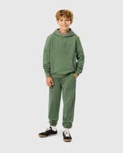 A young person with curly hair stands smiling, wearing the KIDS PACIFIC CHENILLE HOODIE (B0H930D200) and matching joggers from Psycho Bunny. The outfit features a subtle rabbit design, and they complete their stylish look with black sneakers featuring white laces.