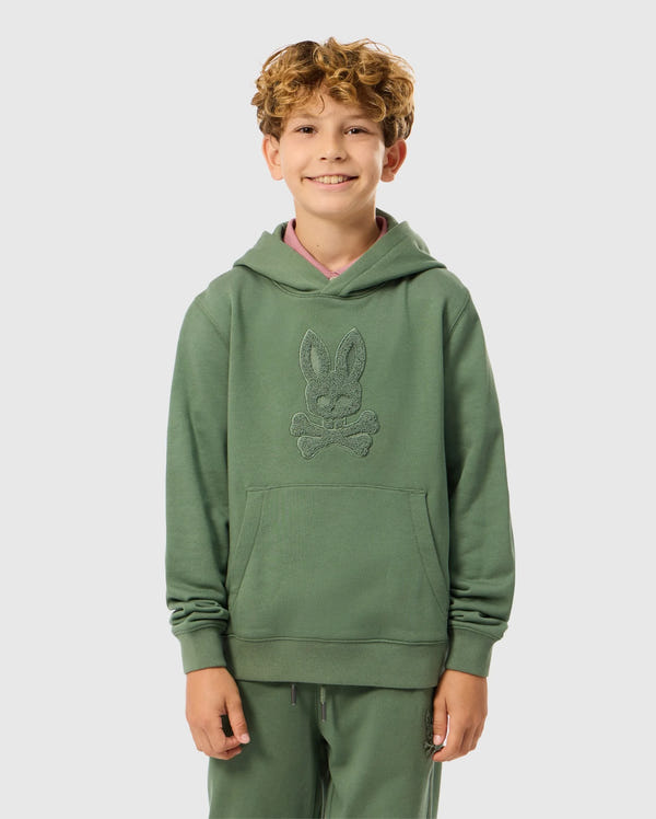 A smiling child with curly hair is wearing the KIDS PACIFIC CHENILLE HOODIE - B0H930D200, by Psycho Bunny. The green French terry hoodie showcases a distinctive design with chenille embroidery. Standing against a plain background, the child has their hands in the front pockets, giving off a casual and comfortable vibe.