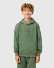 A smiling child with curly hair is wearing the KIDS PACIFIC CHENILLE HOODIE - B0H930D200, by Psycho Bunny. The green French terry hoodie showcases a distinctive design with chenille embroidery. Standing against a plain background, the child has their hands in the front pockets, giving off a casual and comfortable vibe.