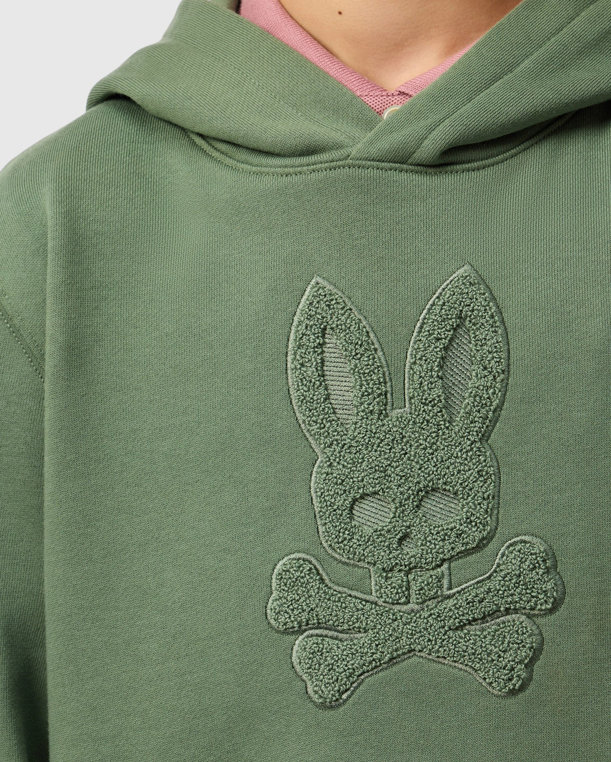 Close-up of a green KIDS PACIFIC CHENILLE HOODIE - B0H930D200 from Psycho Bunny, featuring chenille embroidery of a stylized bunny with long ears above two crossed bones. A hint of a pink shirt is visible underneath the hoodie.